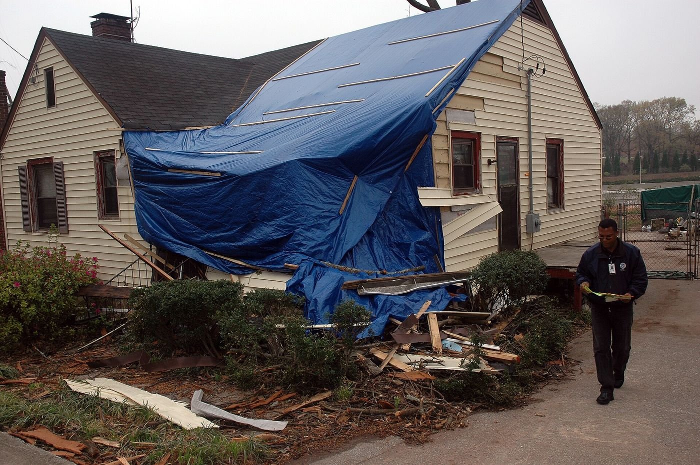 A wind-damaged home with a torn roof and wall covered by a blue tarp, with siding and debris in the yard